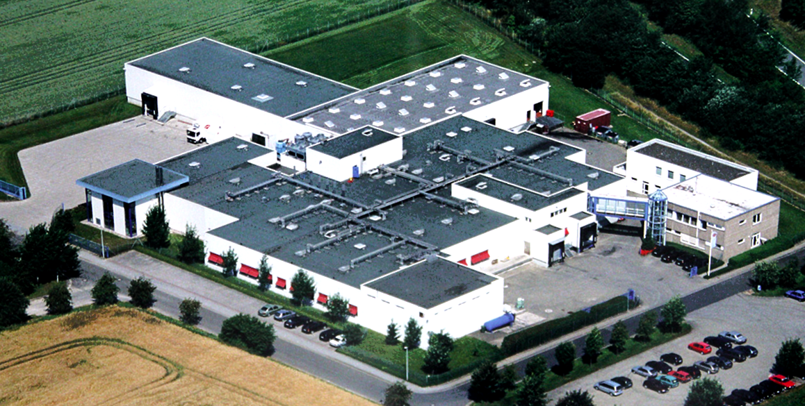 Luftaufnahme eines weißen Industriekomplexes mit mehreren miteinander verbundenen Gebäuden. Parkplätze umgeben den Komplex, im Vordergrund ist ein Feld zu sehen.