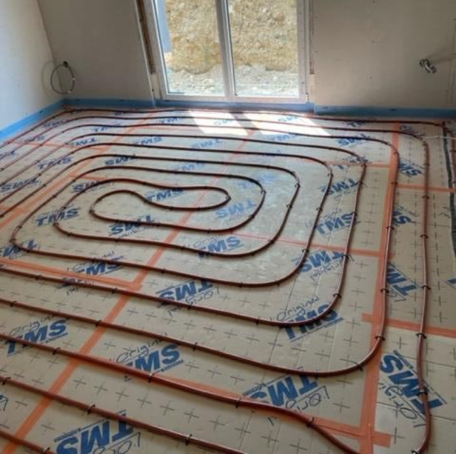 Tuyauterie pour chauffage au sol radiant installée sur une dalle de béton avec des lignes de quadrillage rouges et une isolation périphérique bleue.