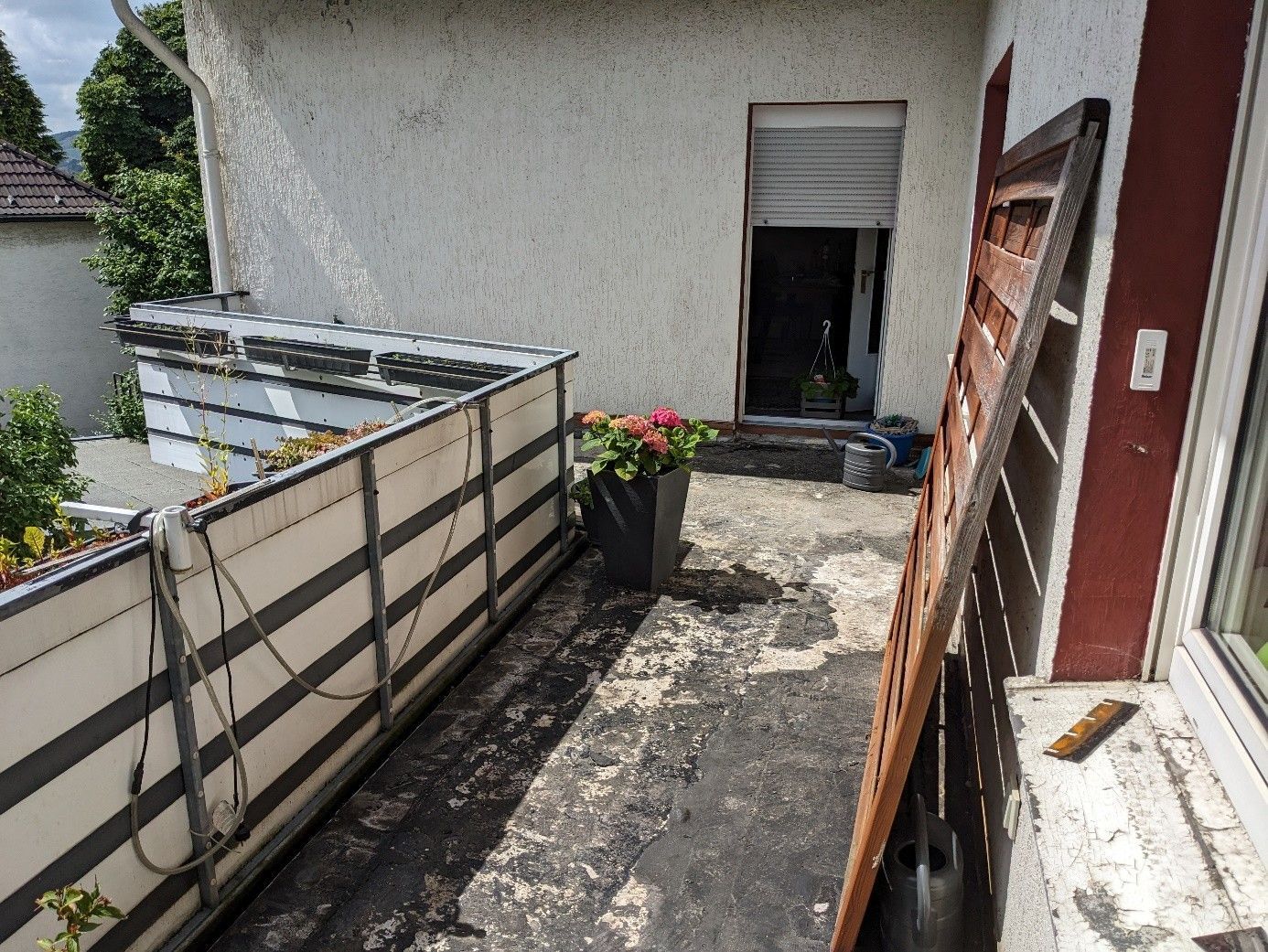 Balkon mit dunkler, abblätternder Oberfläche; geschlossener Fensterladen; Blumentopf; verwitterte Holzkonstruktion.