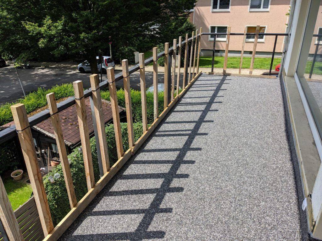 Balkon mit Holzgeländer, grauer Oberfläche und Gebäudehintergrund. Sonnenlicht wirft Schatten auf den Boden.