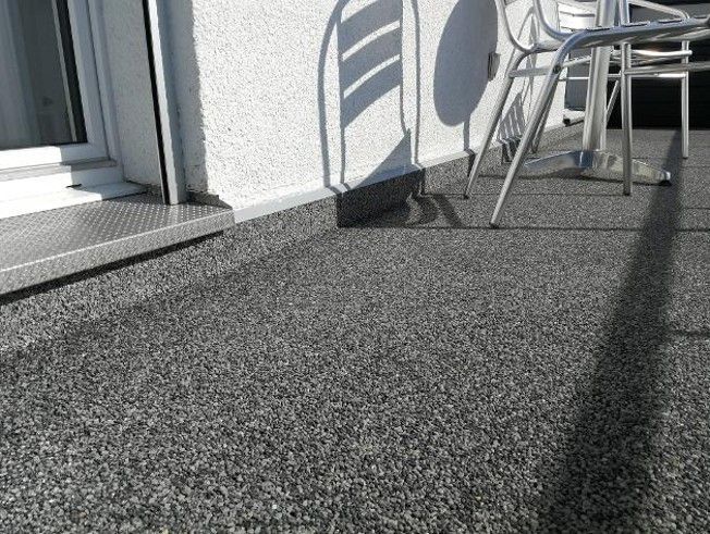 Nahaufnahme eines strukturierten, gesprenkelten grauen Bodens; im Hintergrund neben einem Fenster sind eine weiße Wand und ein Stuhl zu sehen.