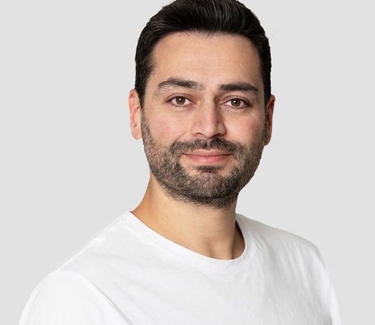 Mann mit dunklem Haar und Bart, trägt ein weißes T-Shirt und lächelt in die Kamera.