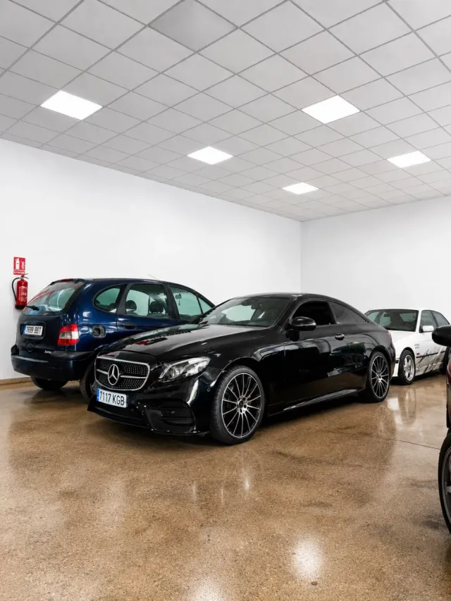 Un sedán Mercedes negro en una sala de exposición interior, con un hatchback azul y un coche blanco cerca.