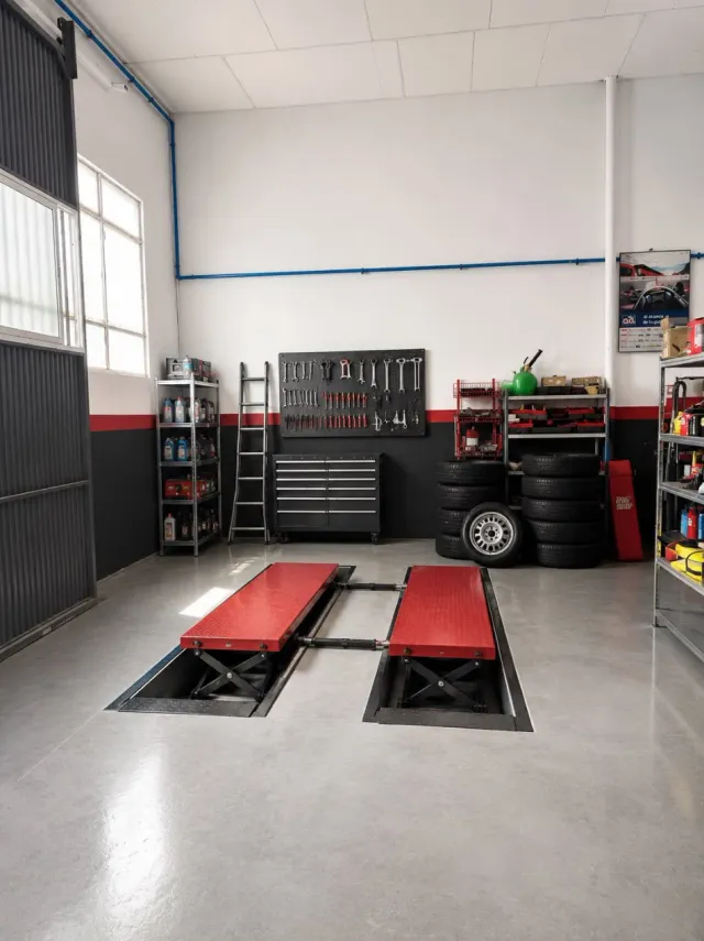 Taller de reparación de automóviles con dos elevadores de vehículos rojos, armarios de herramientas y una pila de neumáticos a lo largo de la pared.