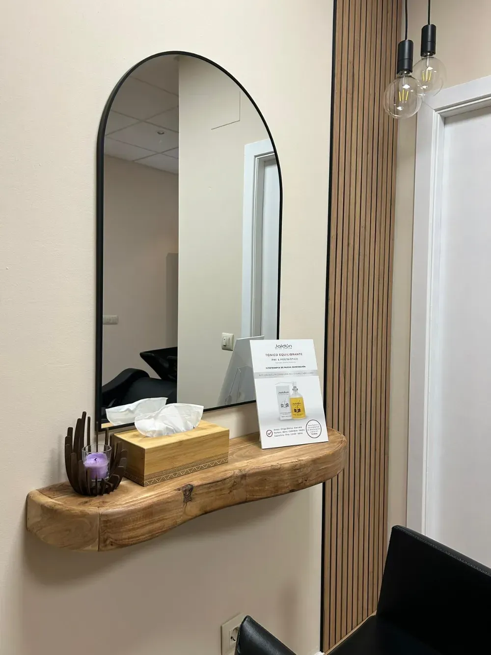 Un espejo arqueado con marco negro cuelga sobre una rústica repisa de madera que sostiene una caja de pañuelos y una vela.