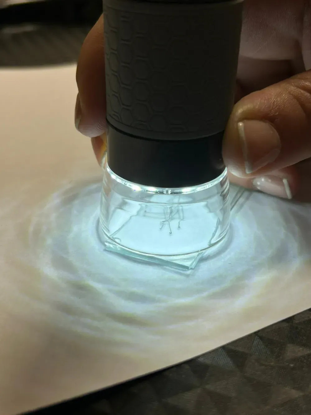 Una persona sostiene un microscopio digital sobre una lámina de vidrio colocada sobre una superficie blanca, iluminada.