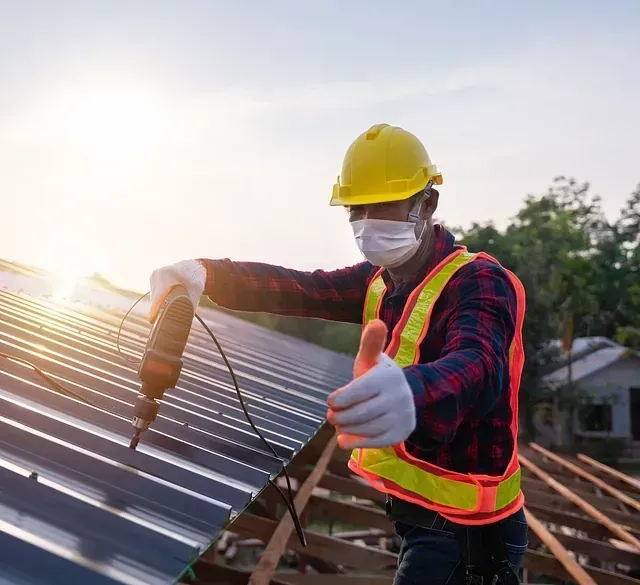 Techador con mascarilla y casco usa un taladro en un techo metálico. Luz solar intensa.