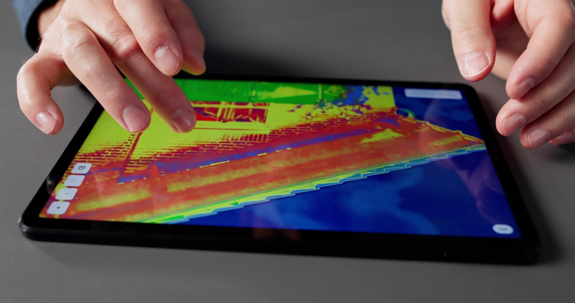 Hände berühren ein Tablet, das ein Wärmebild eines Gebäudes mit farbcodierter Wärmeverteilung anzeigt.