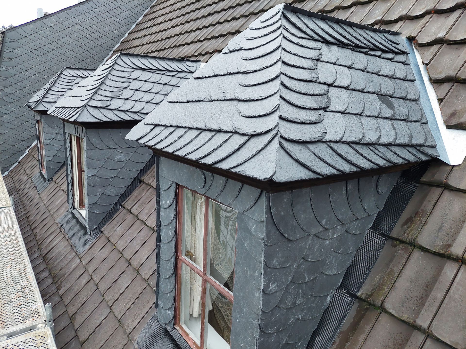 Graue Schiefer-Dachgauben auf einem braunen Schindeldach.