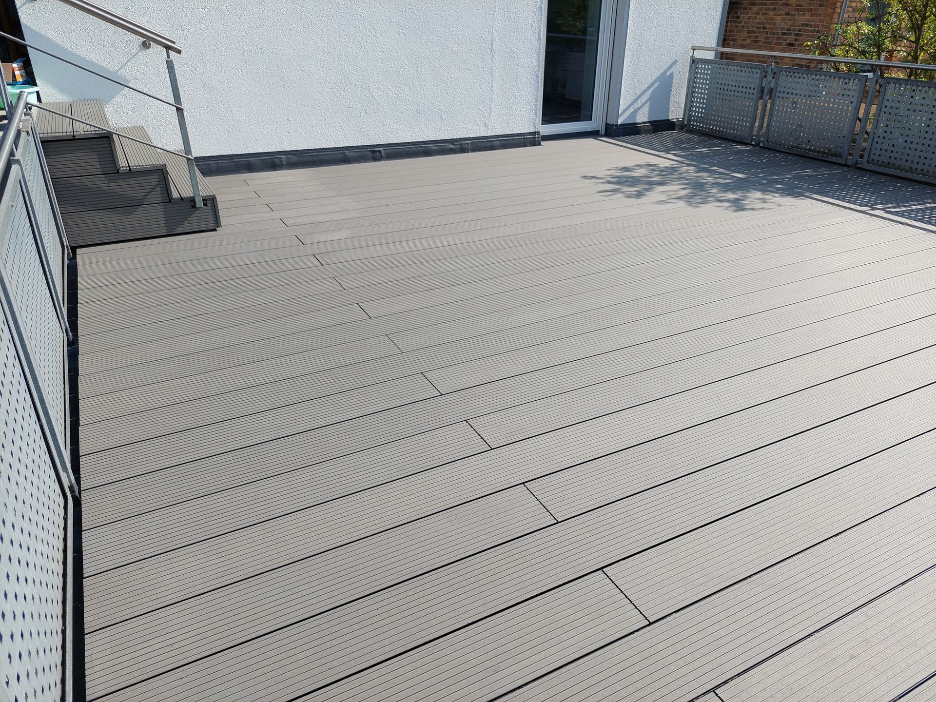 Graue Verbunddielen auf einer Dachterrasse mit Metallgeländer und Treppe. Im Hintergrund ist ein weißes Gebäude zu sehen.