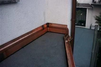 Kupferne Dachrinnen an einer Flachdachecke; weiße Wand, braunes Fallrohr, dunkle Dacheindeckung.