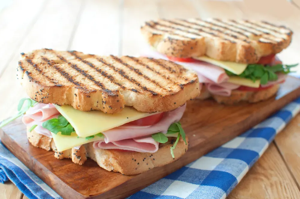 Dos sándwiches a la parrilla con jamón, queso y verduras sobre una tabla de madera, encima de un mantel a cuadros