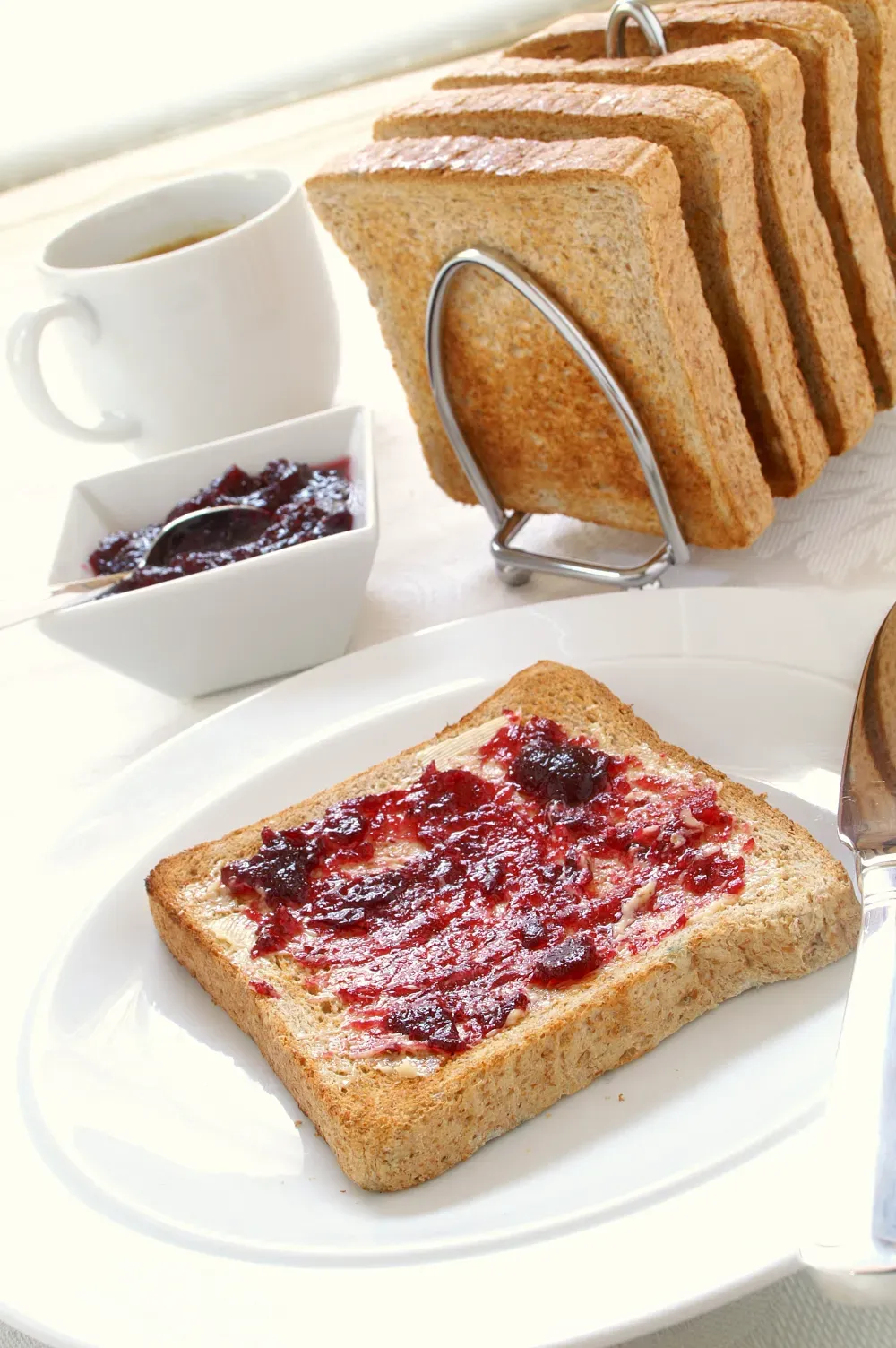 Una rebanada de pan tostado con mermelada en un plato, con una rejilla para tostar, un recipiente para mermelada