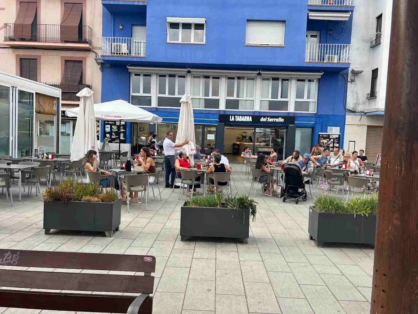 Una cafetería rústica con barra de ladrillo, detalles en verde, mesas, sillas y estanterías repletas de botellas.