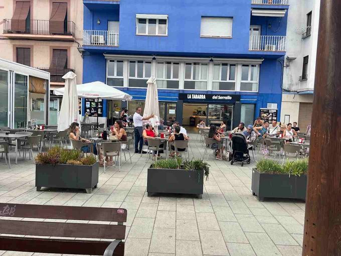 Una cafetería rústica con barra de ladrillo, detalles en verde, mesas, sillas y estanterías repletas de botellas.