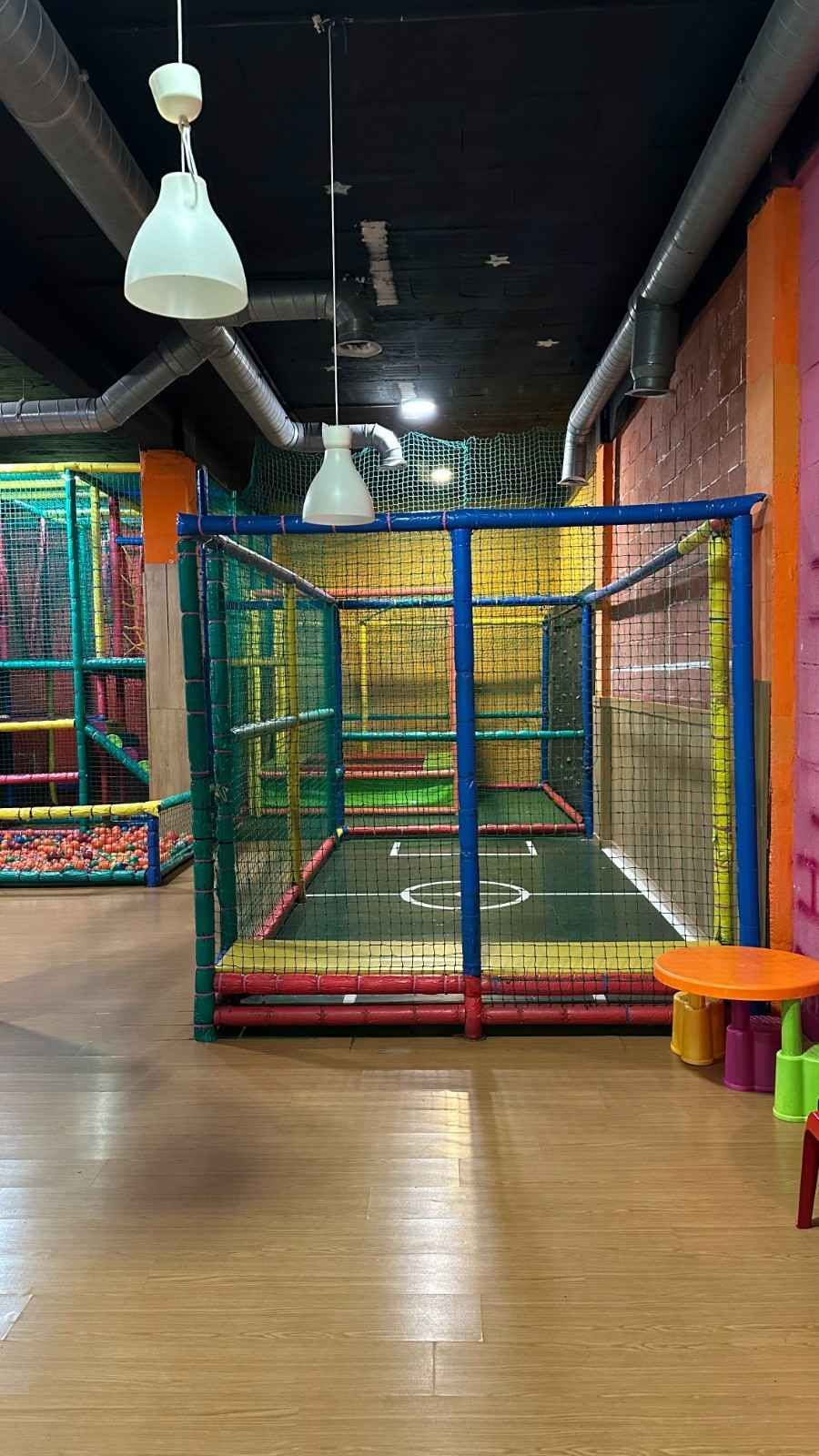 Zona de juegos interior con una colorida jaula de escalada con red, suelo de madera y luces colgantes en forma de cúpula.