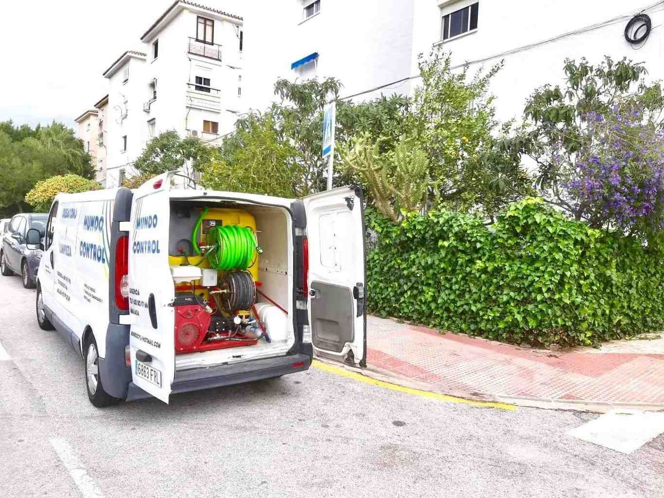 Furgoneta abierta con herramientas aparcada en una calle junto a un seto y un edificio de apartamentos.