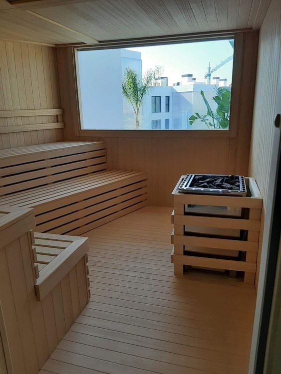 Sauna revestida de madera con bancos y calefacción, con vistas a un luminoso paisaje exterior a través de un gran ventanal.