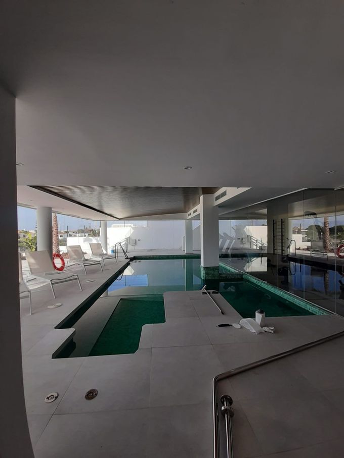Piscina cubierta con agua turquesa, tumbonas y grandes ventanales bajo un techo moderno.