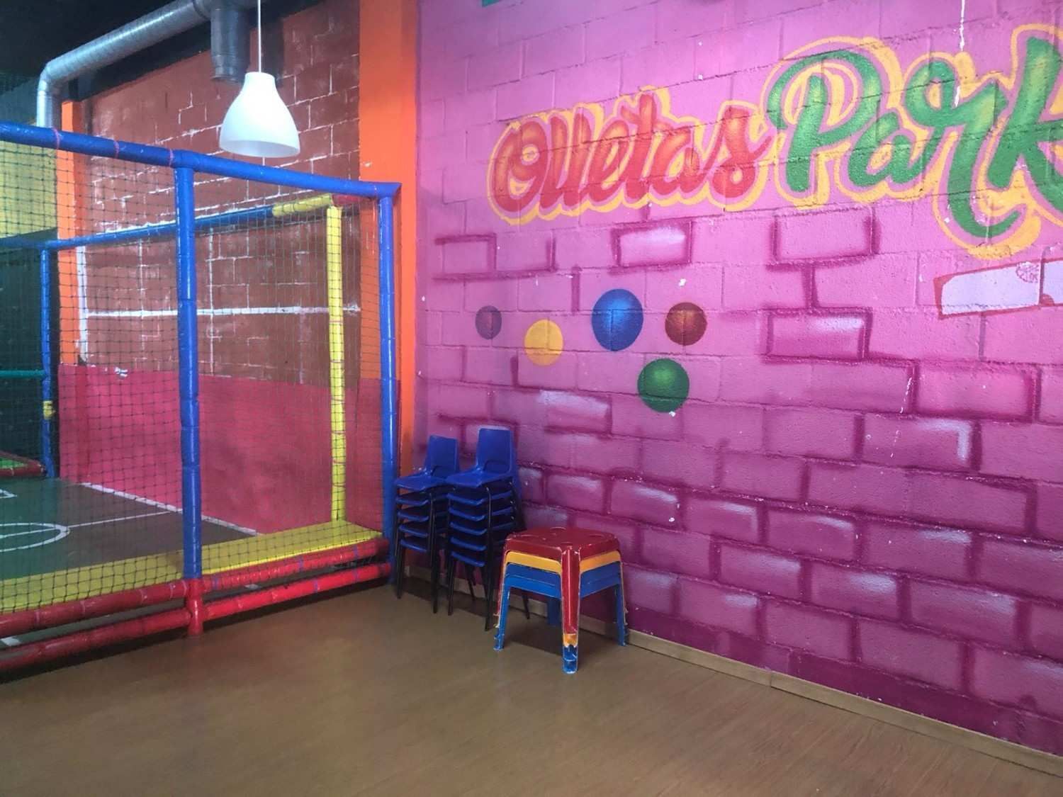 Zona de juegos interior con mural de pared de ladrillo rosa, estructura de escalada con red y sillas coloridas apiladas.