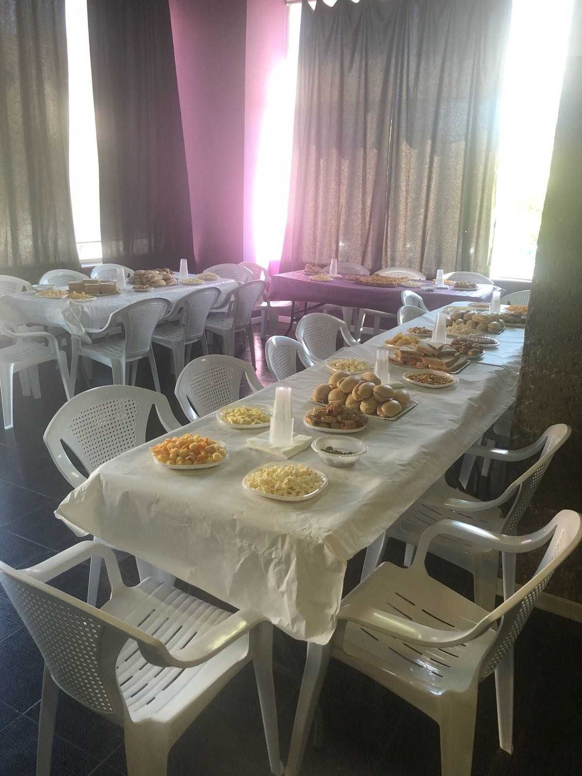 Comedor con una mesa larga puesta con platos de comida y sillas de plástico blancas.
