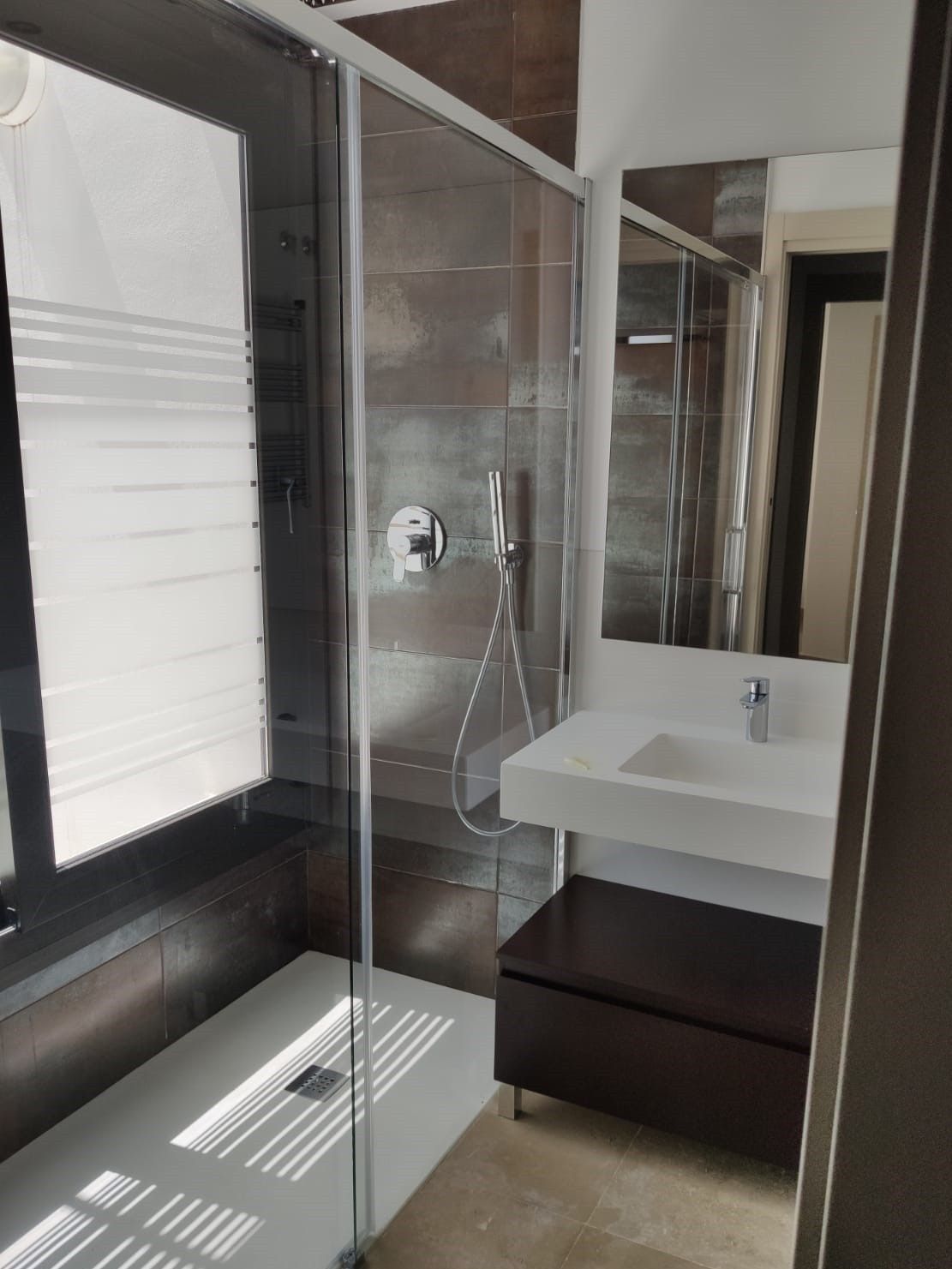 Baño moderno con ducha de cristal, espejo de pared, lavabo blanco y tocador oscuro junto a una ventana luminosa.