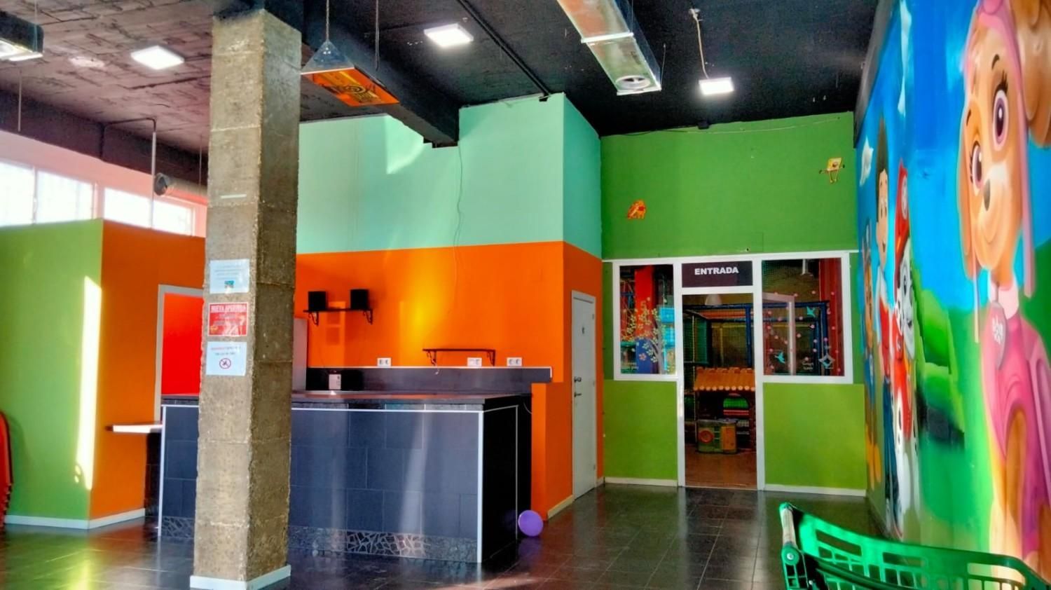 Zona de juegos interior colorida con paredes naranjas y verdes, mostrador, puerta y un gran mural de dibujos animados.