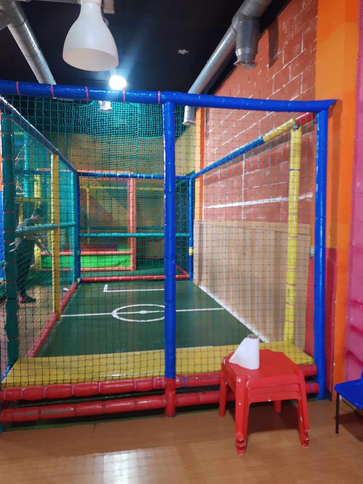 Jaula de fútbol sala con red azul y amarilla, césped verde y un taburete rojo en la parte delantera.