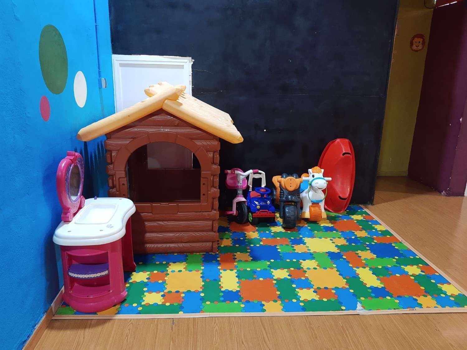 Casita de juegos de juguete y muñecas sobre una colorida alfombra de espuma en una pequeña zona de juegos interior.