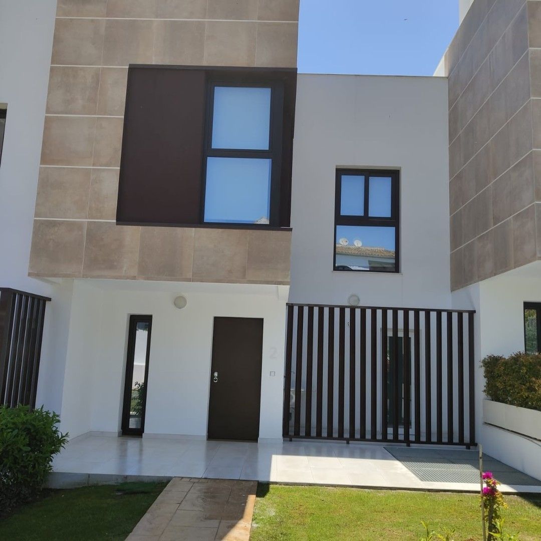 Entrada a una casa adosada moderna con paredes beige, ventanas oscuras, puerta vertical y un pequeño jardín delantero.
