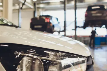 Coche blanco en un taller mecánico con otros dos coches en elevadores; fondo borroso.