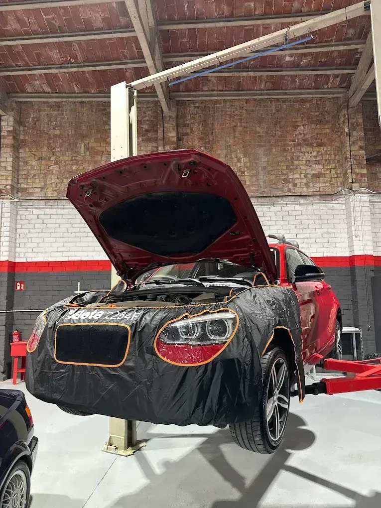 Coche rojo en un elevador en un taller de reparación con el capó abierto, cubierto con una funda protectora negra.