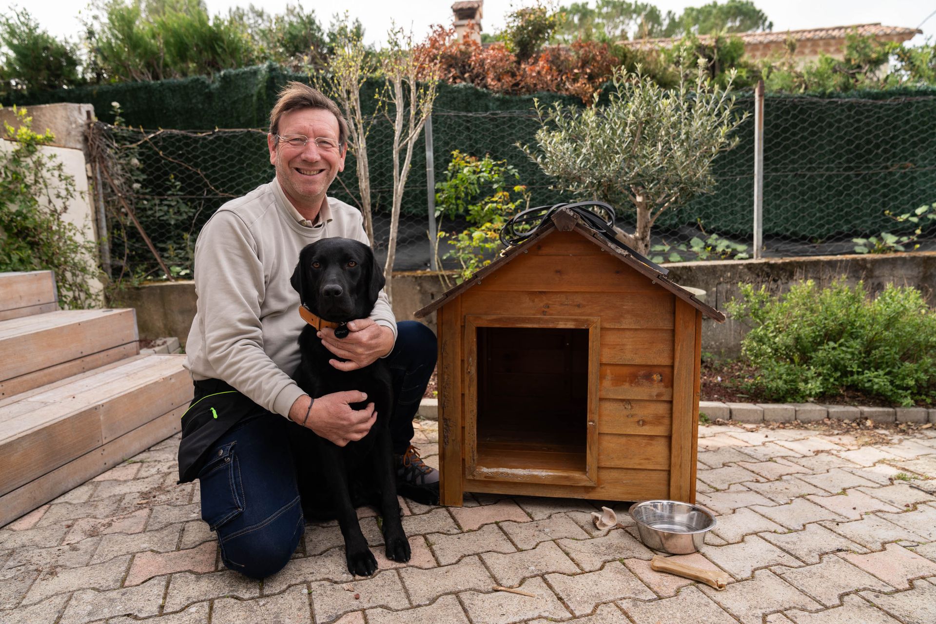 Une personne souriante, agenouillée sur une terrasse en pierre, enlaçant un chien noir à côté d'une niche en bois.