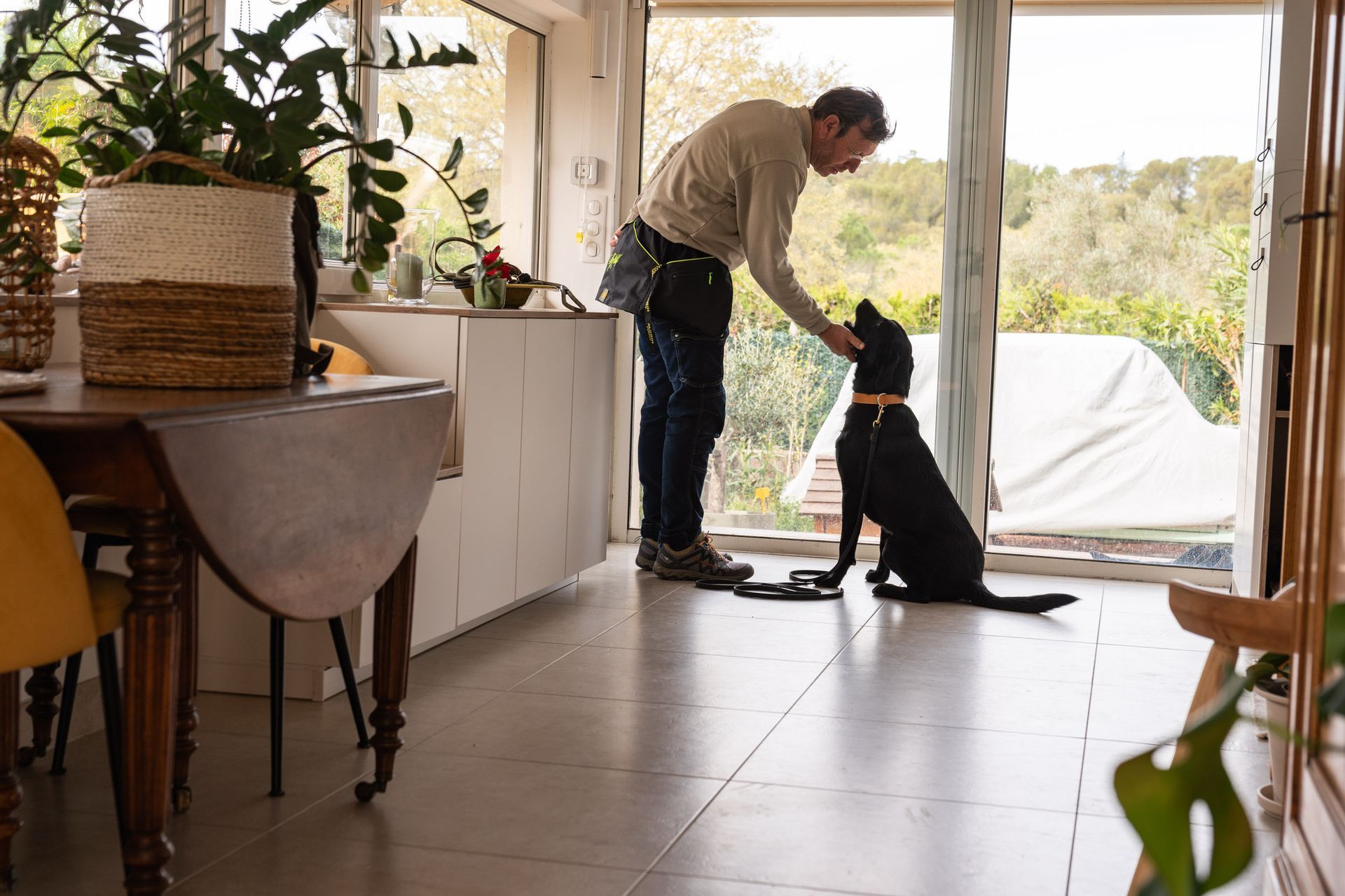 Une personne vêtue d'une chemise claire et d'un jean caresse un chien noir assis sur un sol carrelé dans une pièce ensoleillée.