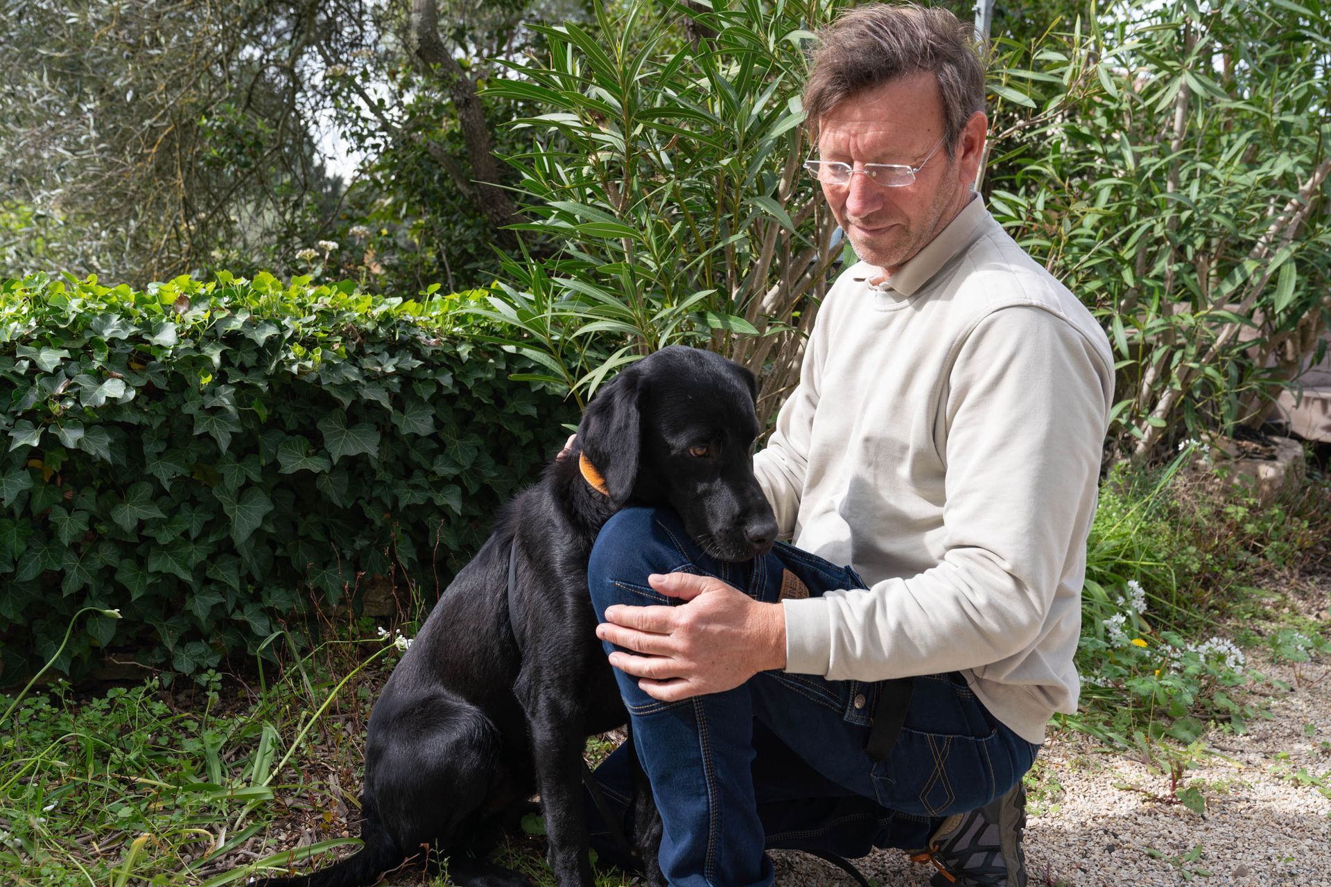 Une personne vêtue d'une chemise beige est agenouillée à l'extérieur, caressant doucement un chien noir portant un collier orange dans un jardin.