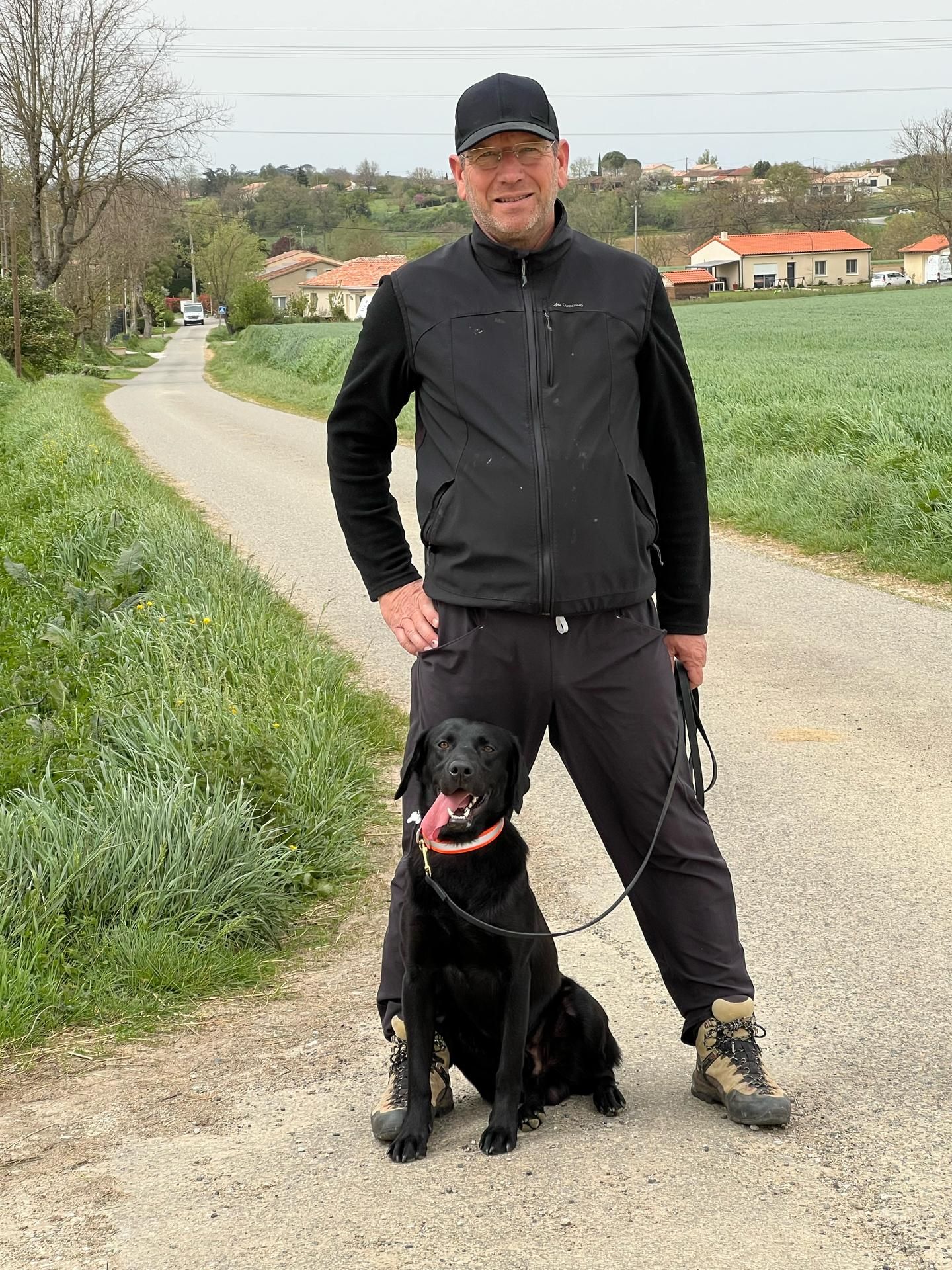 Une personne vêtue d'une veste et d'une casquette noires se tient sur un chemin de gravier, un chien noir assis entre ses jambes.