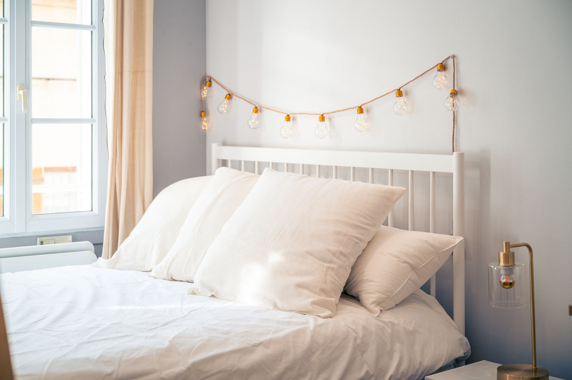 Une chambre confortable avec un lit blanc aux oreillers moelleux et une guirlande lumineuse décorative drapée au-dessus de la tête de lit.
