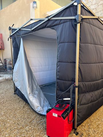 Une tente pliante noire et isolée, installée sur du gravier, avec un chauffage portable rouge fixé à un pied d'angle.