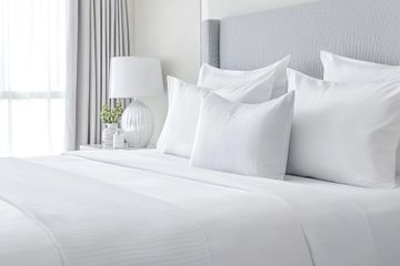 Une chambre lumineuse et propre, avec un lit doté de draps blancs, plusieurs oreillers moelleux et une lampe de chevet.