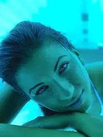 Mujer de piel clara en una cama de bronceado, iluminada por luz azul. Sonríe y mira a la cámara.