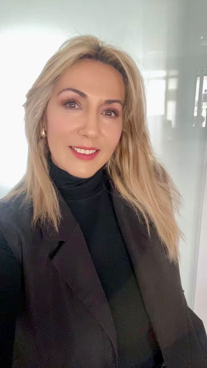 Mujer con cabello rubio, vestida con un jersey de cuello alto y un blazer negro, sonriendo a la cámara en un entorno luminoso.