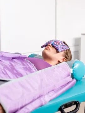 Mujer tumbada en una camilla de masaje, con un antifaz y cubierta con una manta morada. En un ambiente tipo spa.