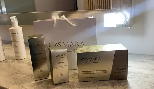 Productos de cuidado de la piel de Casmara en un mostrador, que incluyen una botella, cajas y una bolsa de compras, todo en blanco y plateado.
