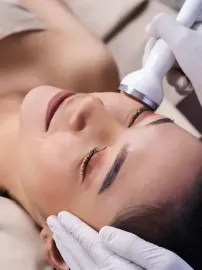 Mujer recibe tratamiento facial para la piel con un dispositivo portátil; manos en guantes blancos.