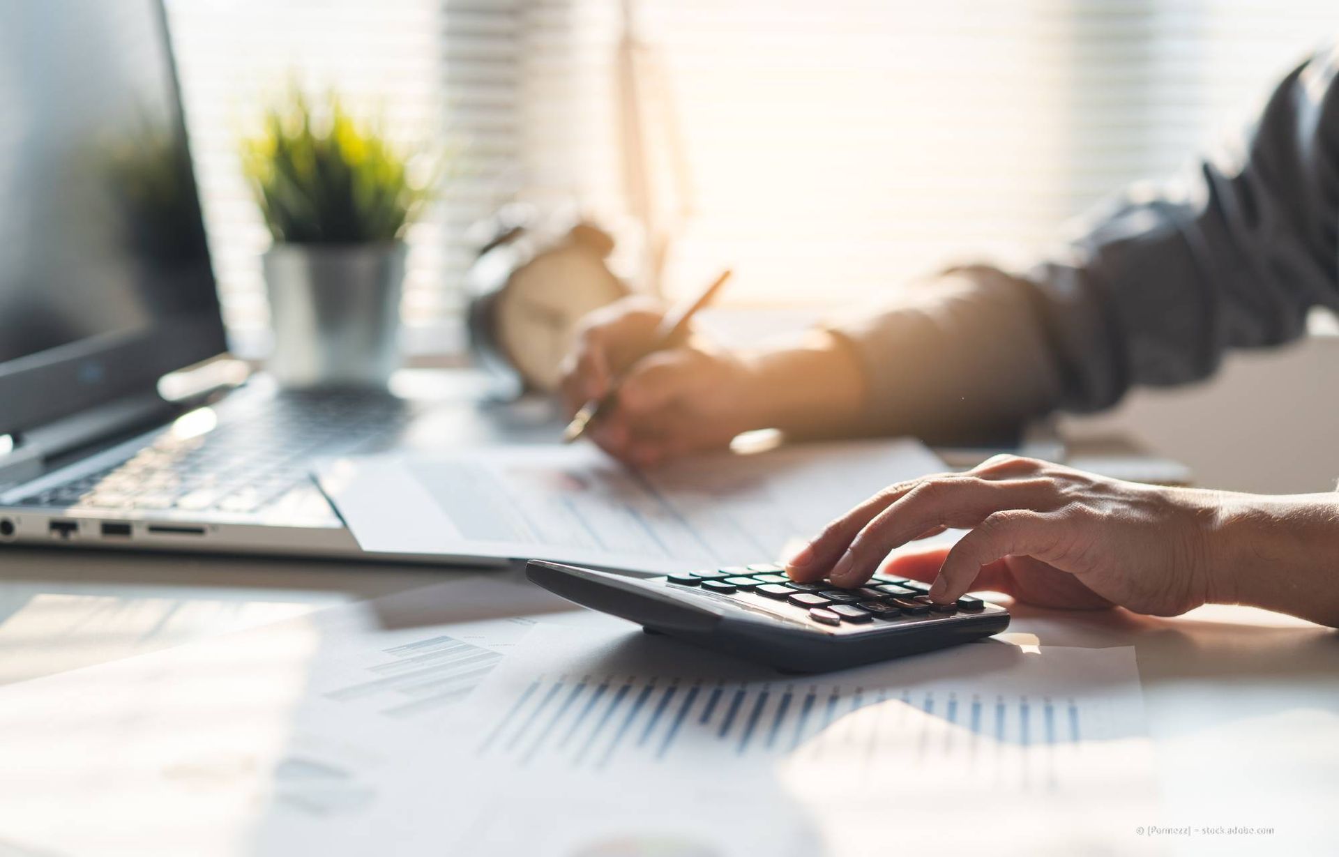 Eine Person sitzt an einem Schreibtisch, neben einem Laptop und einer kleinen Pflanze, und benutzt einen Taschenrechner und schreibt auf Papier.