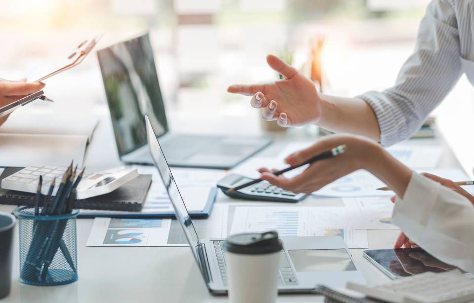 Menschen arbeiten gemeinsam an einem Schreibtisch, benutzen Laptops, Papier und Taschenrechner und besprechen ihre Arbeit.