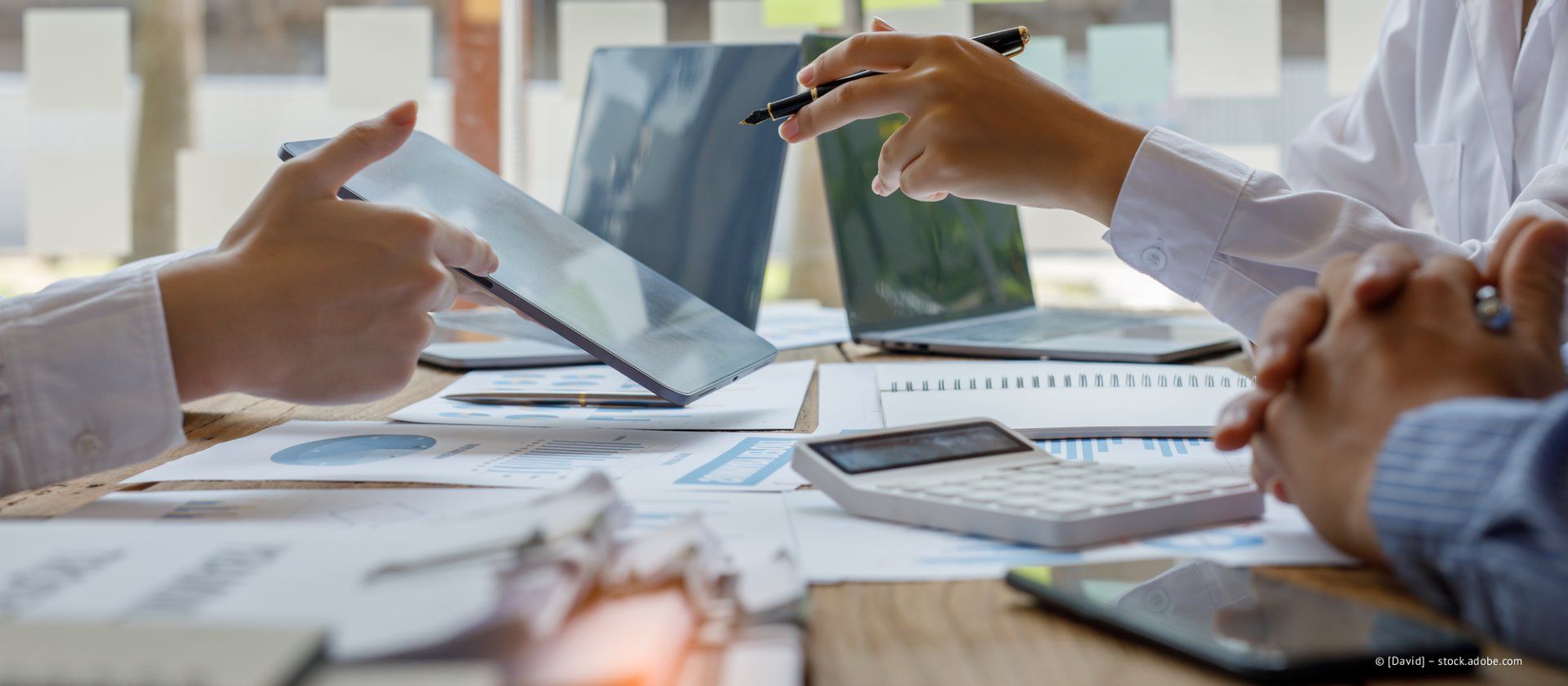 Eine Hand hält einen silbernen Stift und schreibt auf ein Dokument mit blauen Diagrammen. Im Hintergrund ist ein Laptop zu sehen.