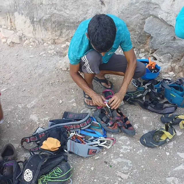 Un hombre está arrodillado frente a una pila de equipo de escalada, incluido un par de sandalias con la letra s.