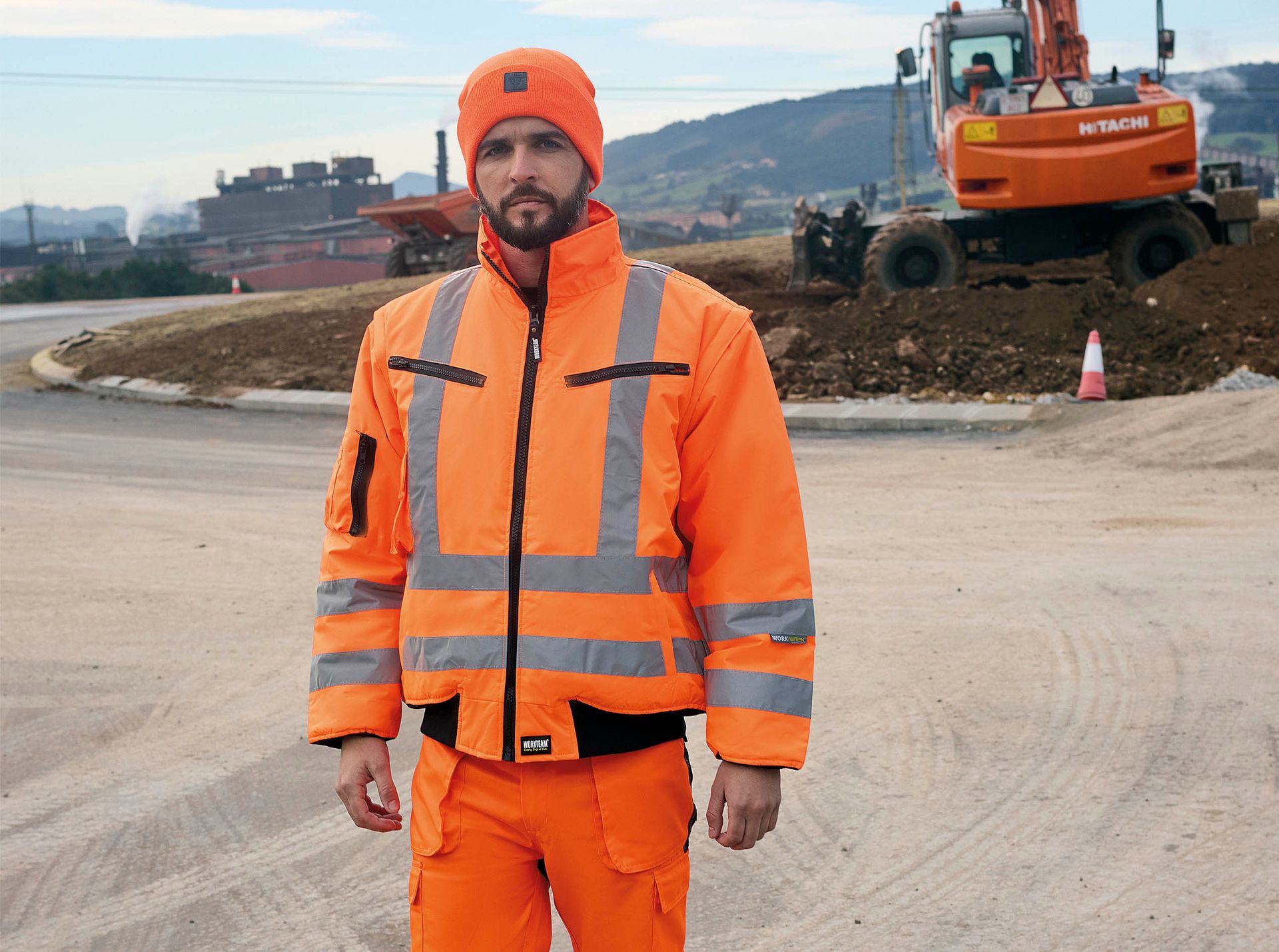 Un hombre que viste una chaqueta naranja y pantalones naranjas está parado frente a una excavadora.
