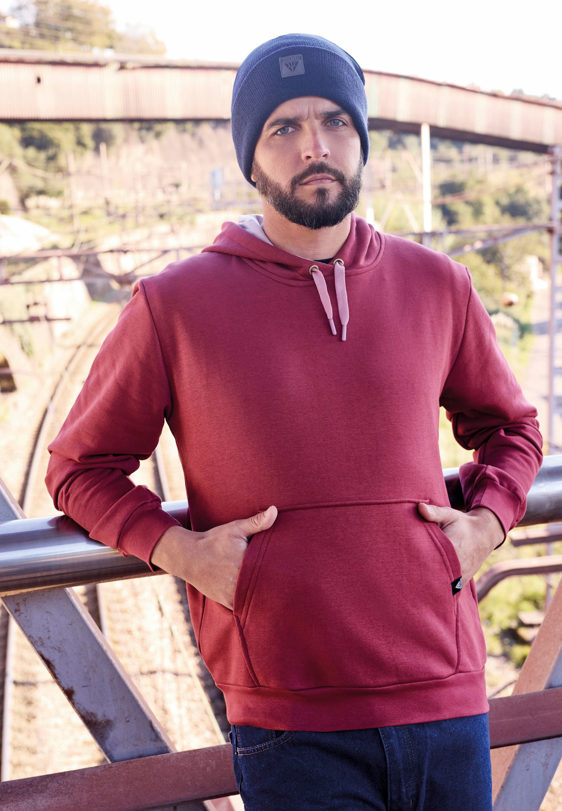 Un hombre que viste una sudadera con capucha marrón y un gorro negro está de pie junto a una barandilla.